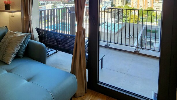 Una sala de estar moderna con un sofá, un ventanal con cortinas y vistas a un paisaje urbano y edificios.