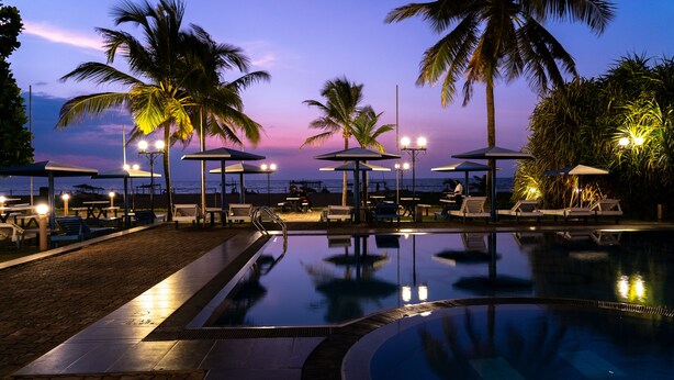 A pool area with lounge chairs and umbrellas, a beach view, and a sunset. - Rani Beach Hotel (Negombo)