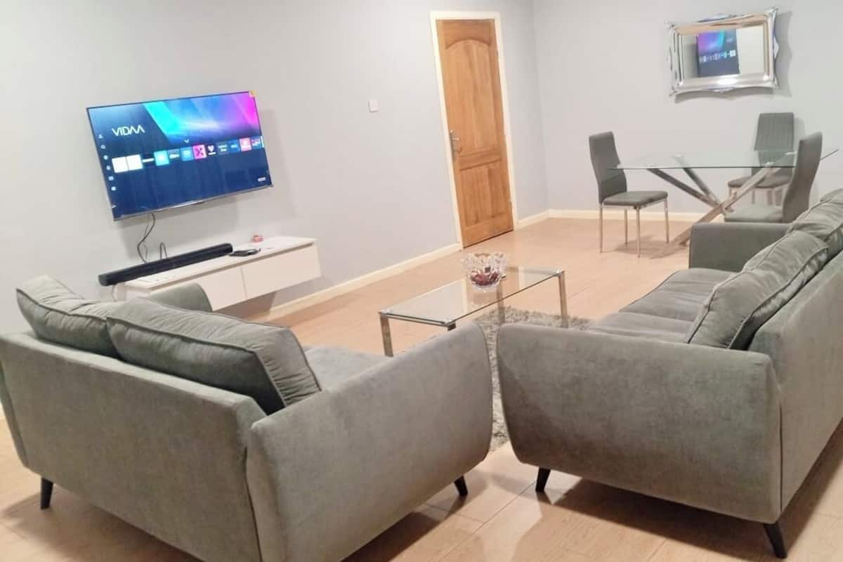 A modern living room with a grey sofa set, a flat-screen TV mounted on the wall, a glass coffee table, and a dining area with a glass table and chairs.