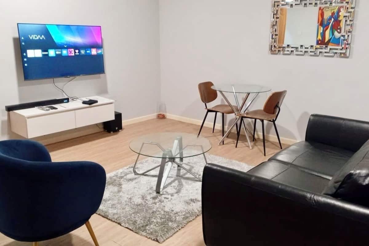 A modern living room with a flat-screen TV, a glass coffee table, a black leather sofa, and two brown chairs.