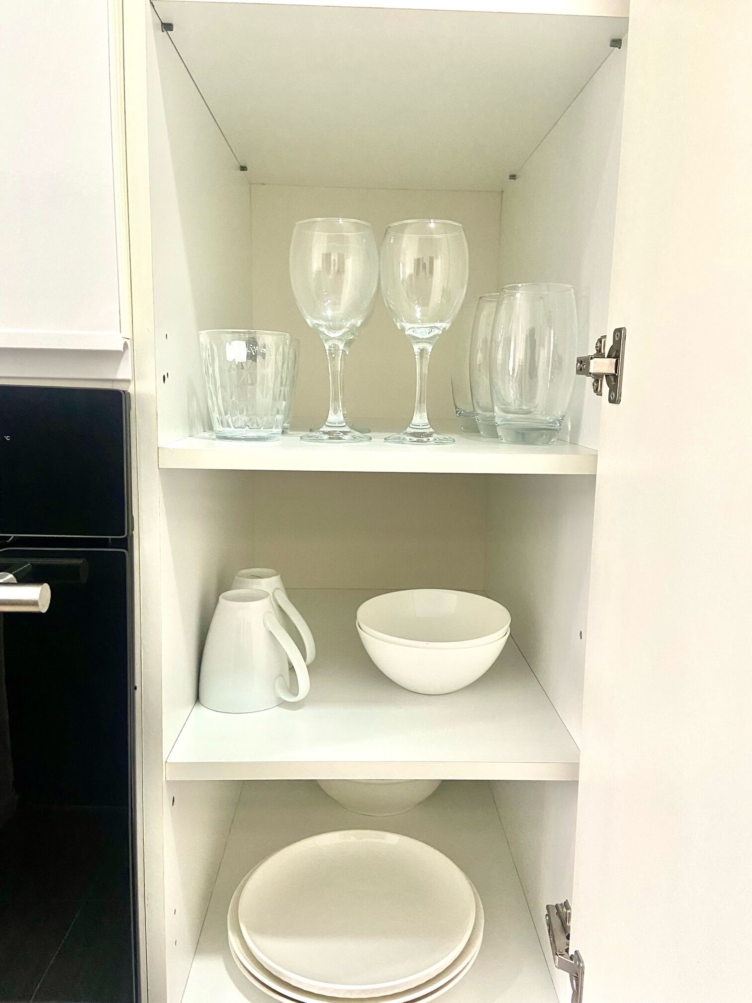 A kitchen cabinet with shelves holding various glassware, a white pitcher, and stacked white plates.