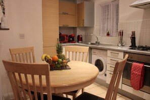 A compact kitchen with wooden cabinets, a round dining table, and a washing machine. - Apartment - Sleeps 2 - Parking - Station Close (Dagenham)