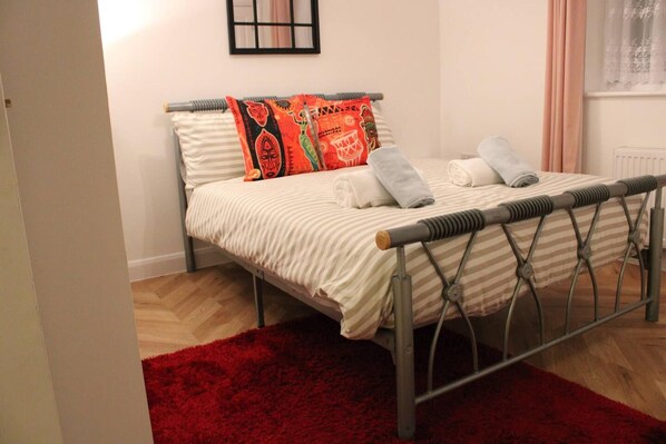 A neatly made bed with striped bedding and decorative pillows, a red rug, and a metal headboard. - Apartment - Sleeps 2 - Parking - Station Close (Dagenham)