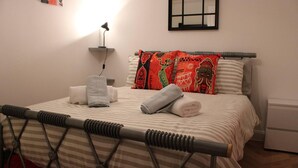 A neatly made bed with striped bedding, a headboard, and decorative pillows. There is a bedside lamp, a small shelf with a lamp, and a wall-mounted mirror. - Apartment - Sleeps 2 - Parking - Station Close (Dagenham)