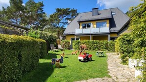 A house with a stone pathway leading to a lawn with two toy cars and a swing set. - Fewo 4 Triftweg 10 (Loddin)