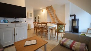 A cozy living room with a sofa, a wooden dining table with chairs, a television on a cabinet, a staircase, and a fireplace. - Fewo 4 Triftweg 10 (Loddin)