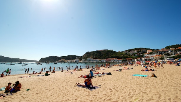 A crowded beach with people sunbathing and swimming, surrounded by buildings and boats.