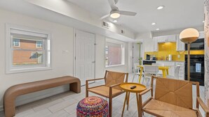 A modern kitchen with white cabinets, a yellow backsplash, and a black oven. There is a dining area with a bench and two chairs, and a colorful ottoman. - TreeHouse Lodge*Walk to Beach*Pet-Friendly (#106) (North Myrtle Beach)
