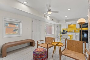 A modern kitchen with white cabinets, a yellow backsplash, and a black oven. There is a dining area with a bench and two chairs, and a colorful ottoman.