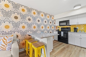 A modern kitchen with white cabinets, a black microwave, and a yellow backsplash. There are two yellow stools and a white countertop.
