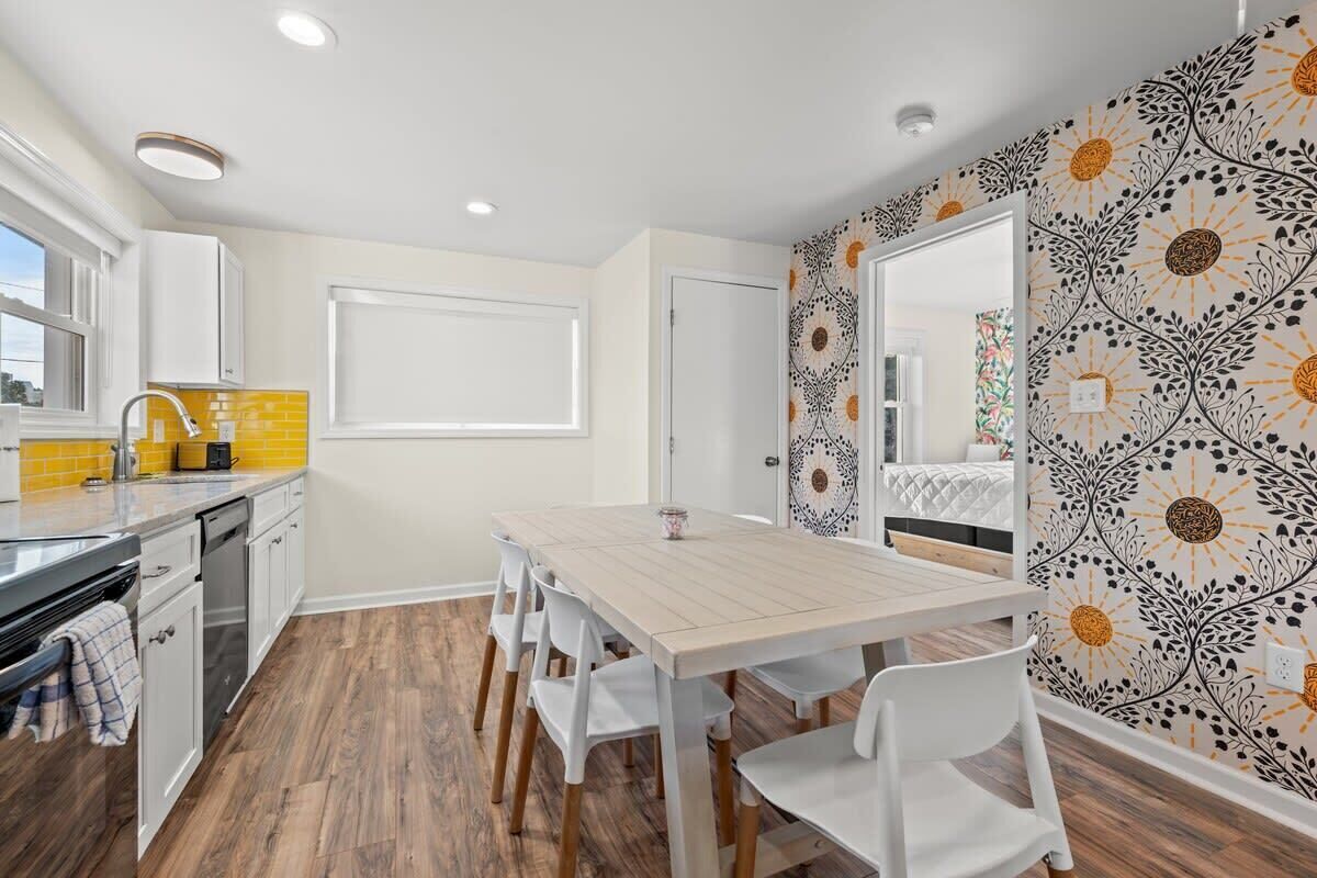 A compact kitchen with a wooden table and white chairs, a yellow backsplash, and a patterned wall.