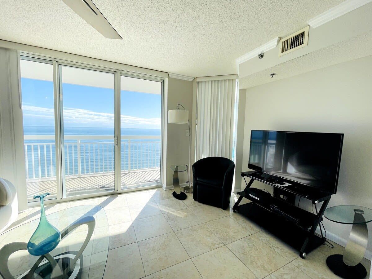 A spacious room with a large window offering a view of the ocean, a flat-screen TV, a glass coffee table, and a black chair.