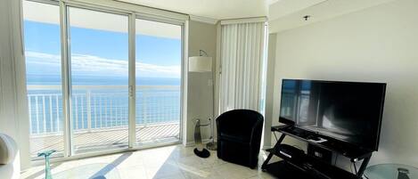 A spacious room with a large window offering a view of the ocean, a flat-screen TV, a glass coffee table, and a black chair.