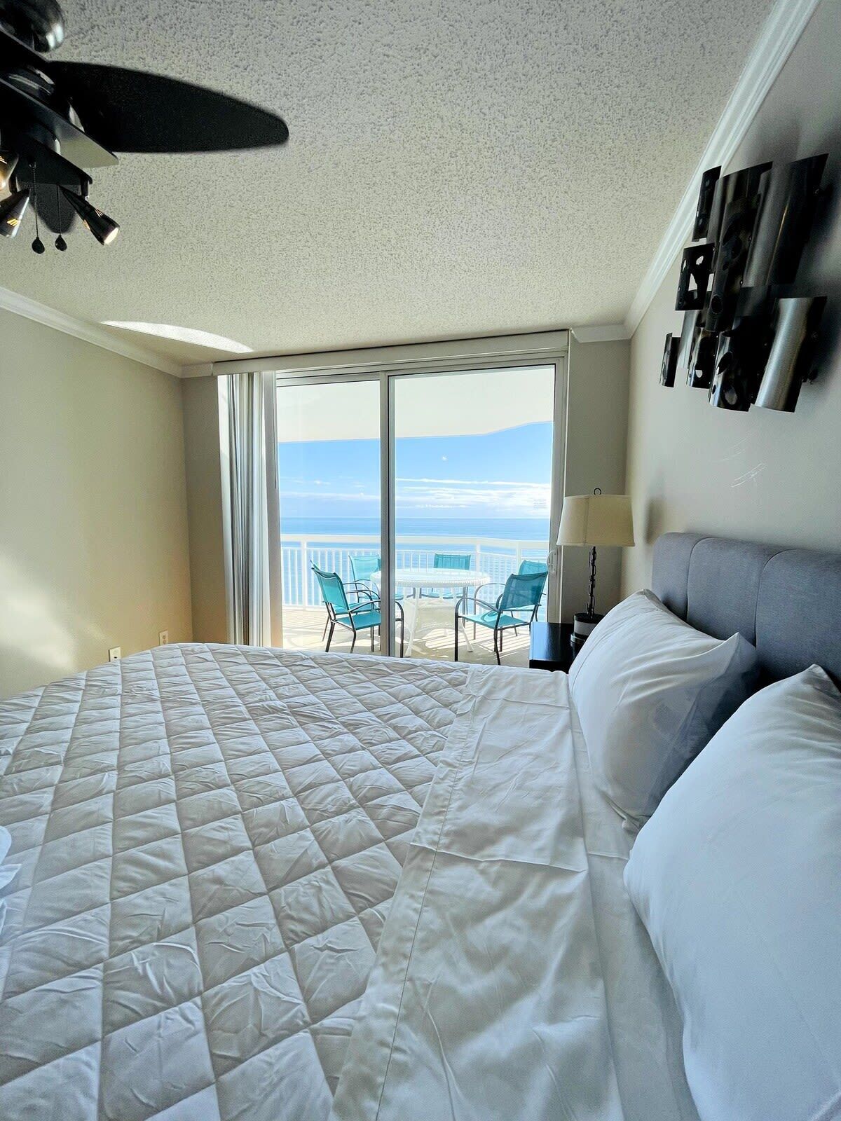 A hotel room with a large bed, a ceiling fan, a balcony with chairs, and a view of the ocean.