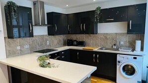 A modern kitchen with dark cabinets, a central island, a built-in washing machine, and wooden flooring. - Modern City Center 2 Bedroom Apartment (Aberdeen)
