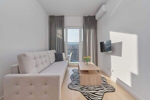 A modern living room with a sofa, a coffee table, a zebra-patterned rug, a TV mounted on the wall, and a view of the city through large windows.