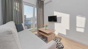 A modern living room with a sofa, a coffee table, a TV, and a view of buildings through large windows. - Modern Apartment | Parking, Balcony, Workspace (Wroclaw)