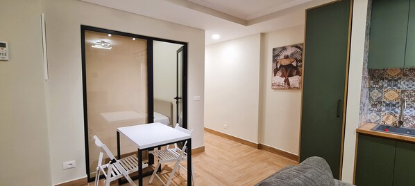 Un espacio habitable pequeño y de planta abierta con kitchenette, un comedor con mesa y sillas blancas, y un espejo grande.