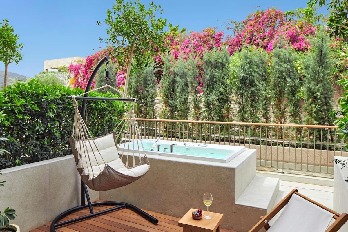 Um deck de madeira com uma cadeira suspensa, uma mesa pequena com uma taça de vinho, e vista para uma piscina e vegetação exuberante.