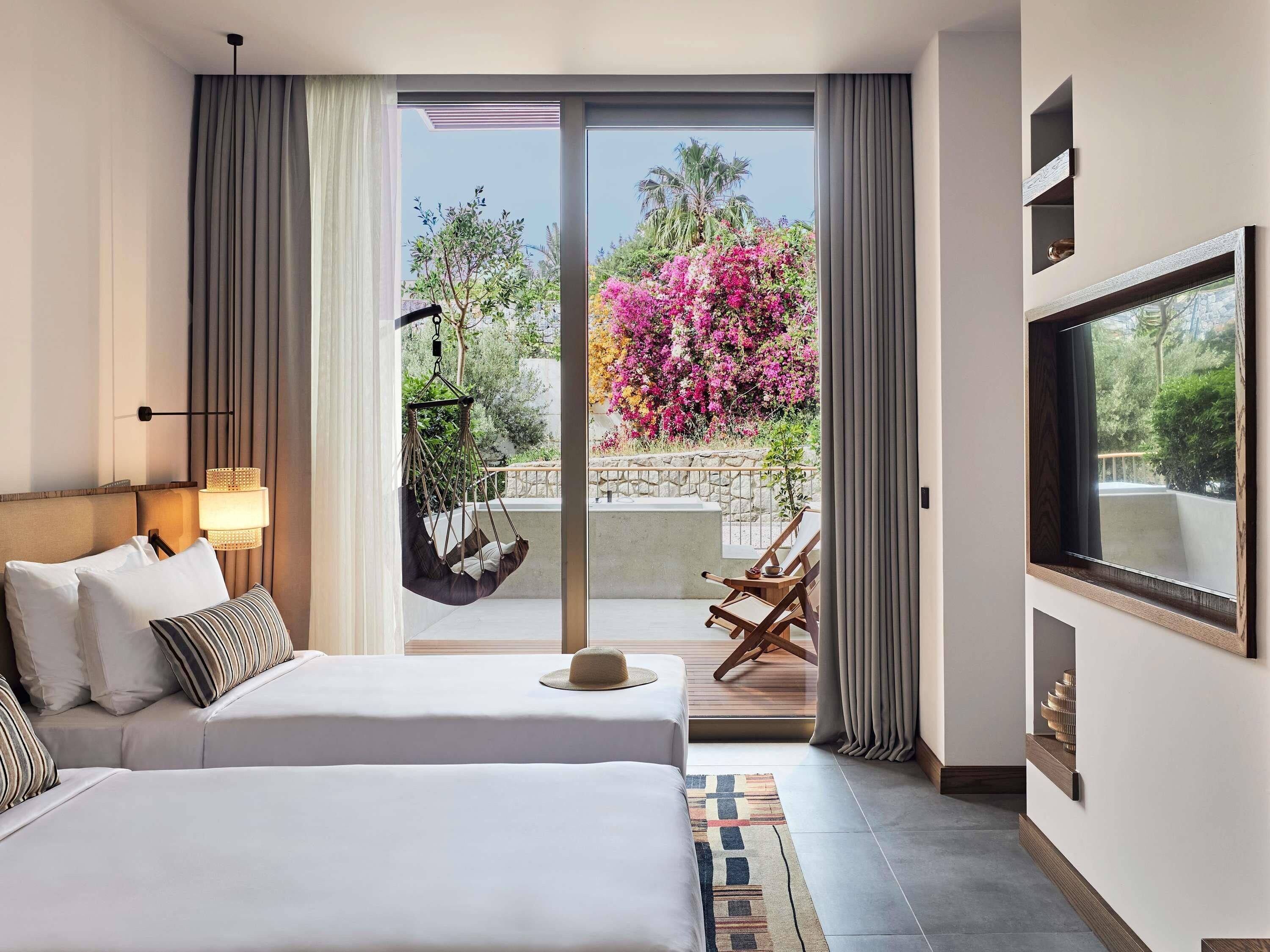 Ein Hotelzimmer mit einem großen Bett, einem Hängesessel, einem Fernseher und einem Balkon mit Blick auf Bäume und Blumen.