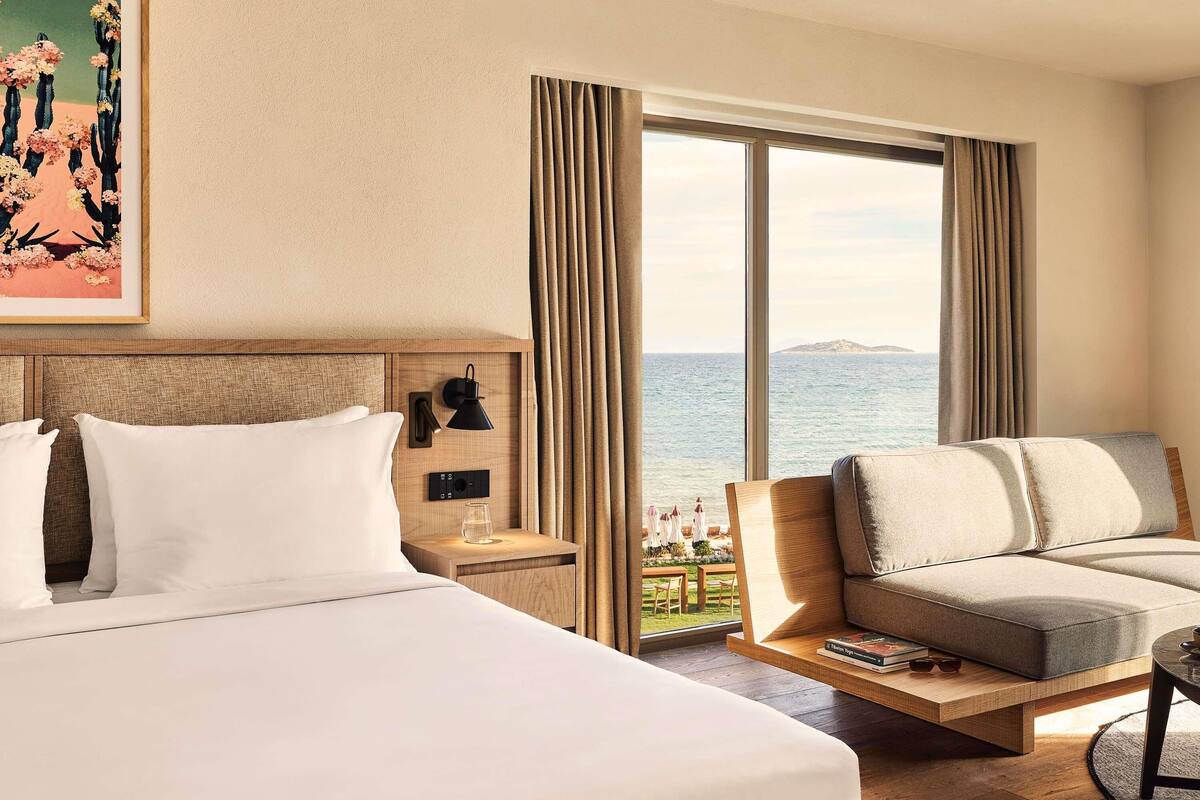 Quarto de hotel com uma cama, área de estar com vista para o mar, mesa de cabeceira com abajur e um quadro na parede.