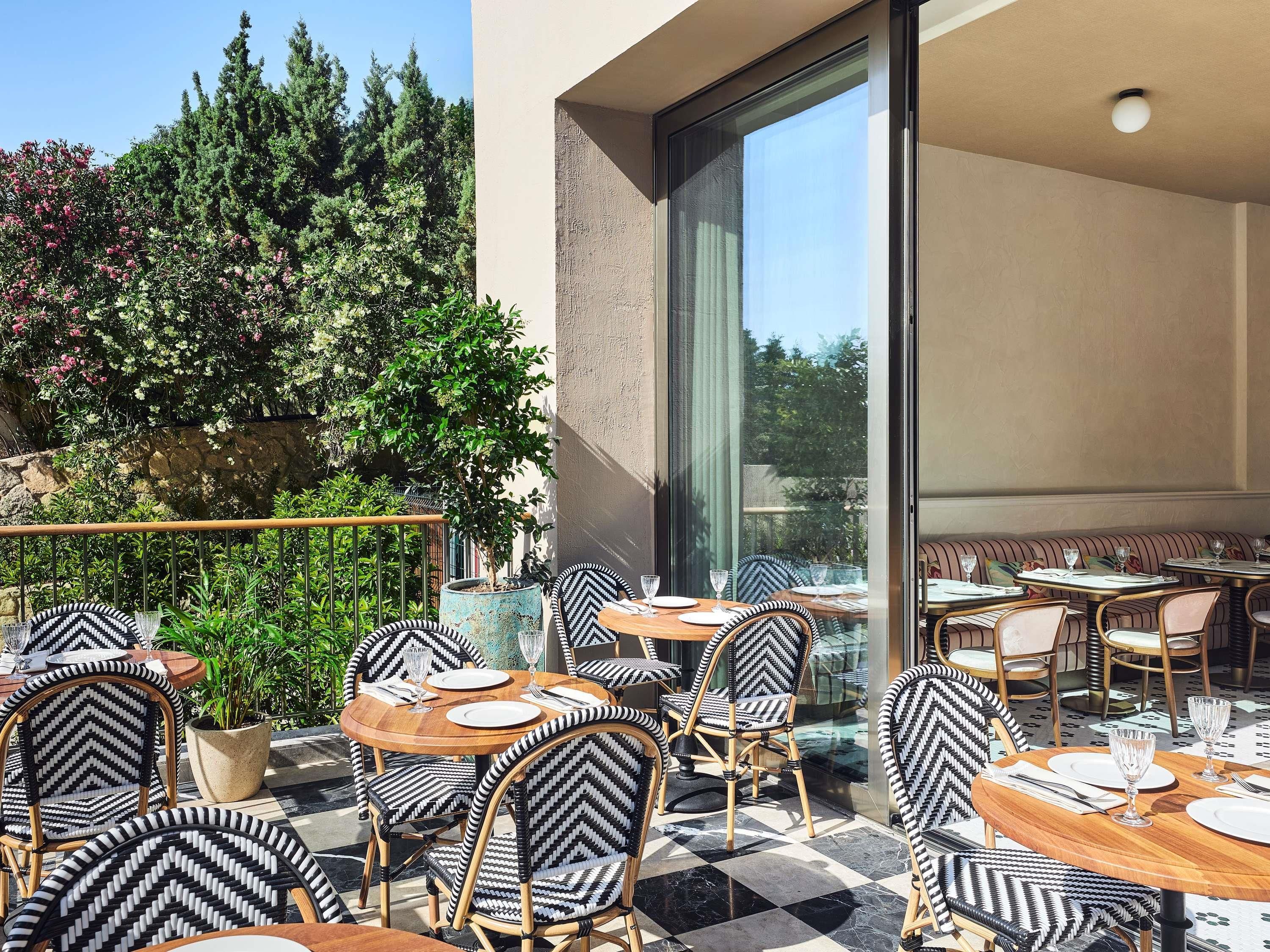 An outdoor dining area with round tables and chairs, a view of greenery, and a large window.