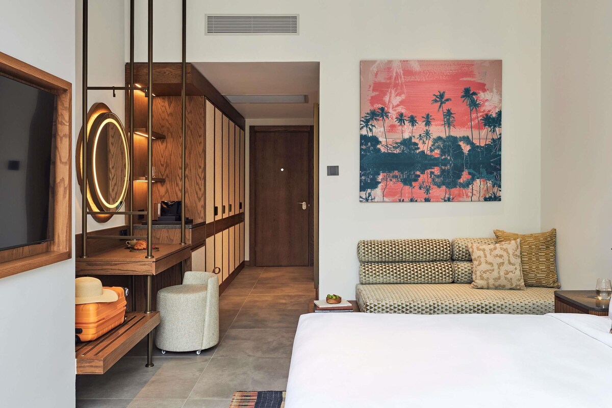 Une chambre d’hôtel moderne comprenant un lit, un canapé, une étagère en bois où sont posés des bagages, un téléviseur fixé au mur et un tableau sur le thème des pays tropicaux.