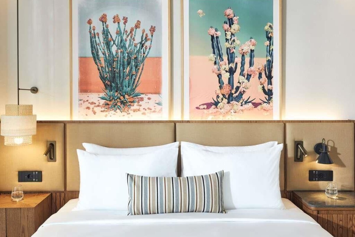 A hotel room with a bed, two bedside tables, a lamp, and two framed artworks on the wall.
