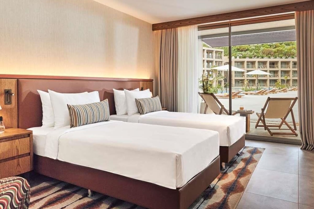 A hotel room with two beds, a large window with curtains, a view of the hotel complex, and a small balcony area.