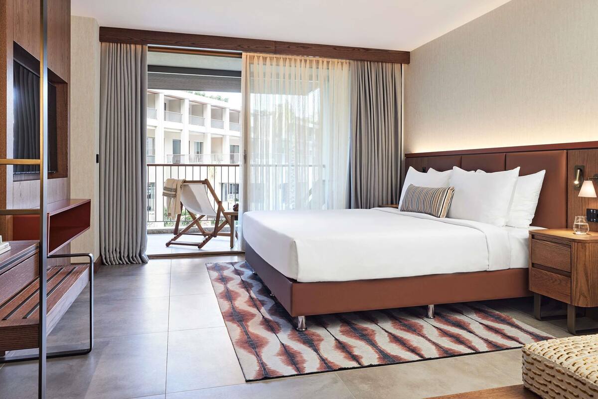 Quarto de hotel com uma cama grande, mesa de cabeceira de madeira, escrivaninha, cadeira e vista para um edifício pela janela.