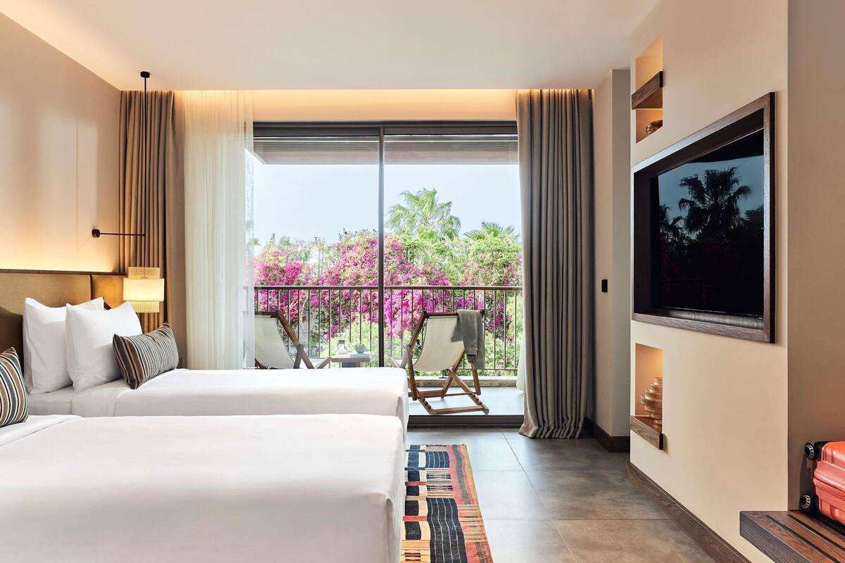 Quarto de hotel com cama, cadeira, televisão e varanda com vista para flores cor-de-rosa.