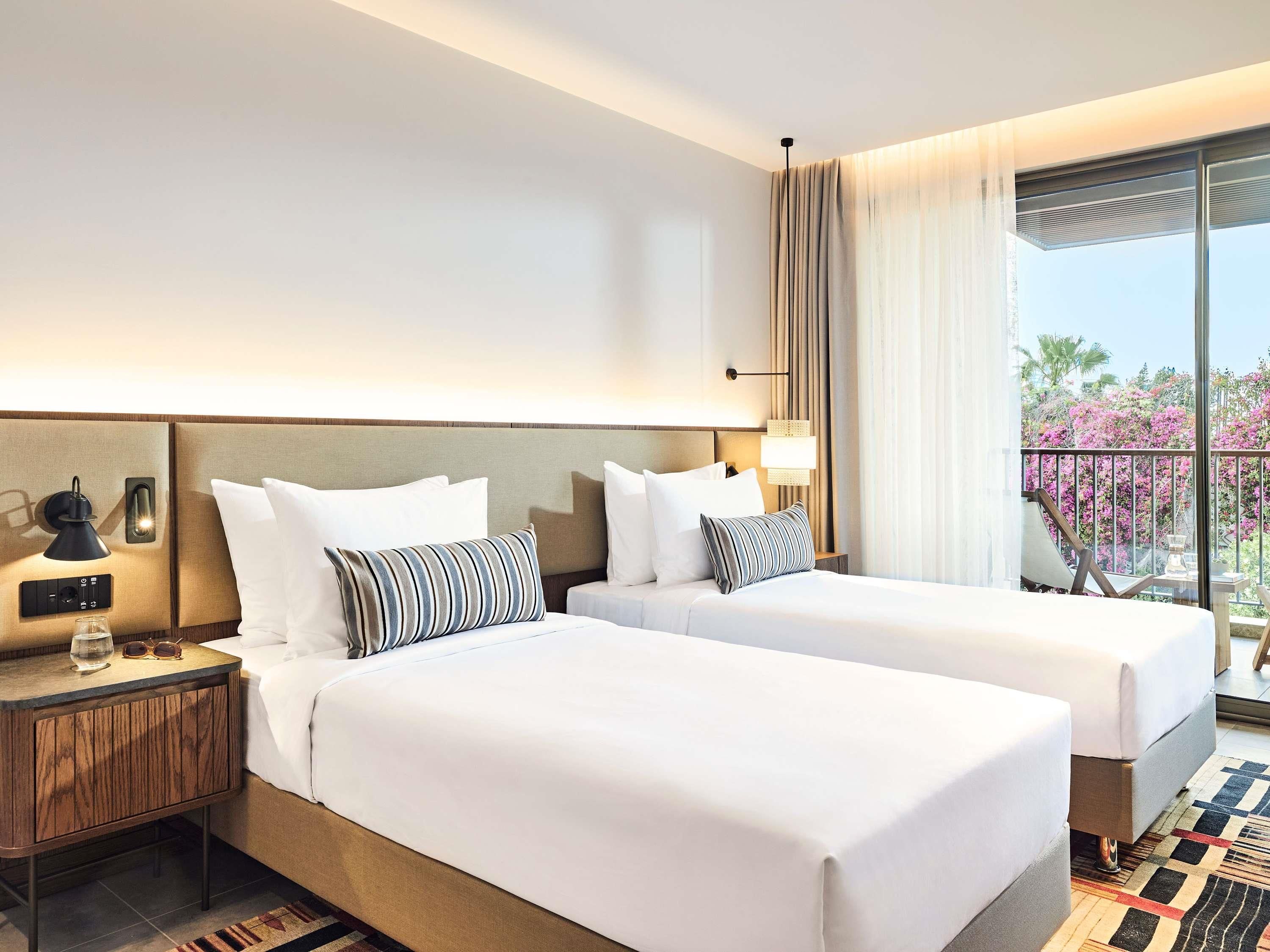 A hotel room with two beds, a nightstand with a lamp, a window with curtains, and a view of the outdoors.