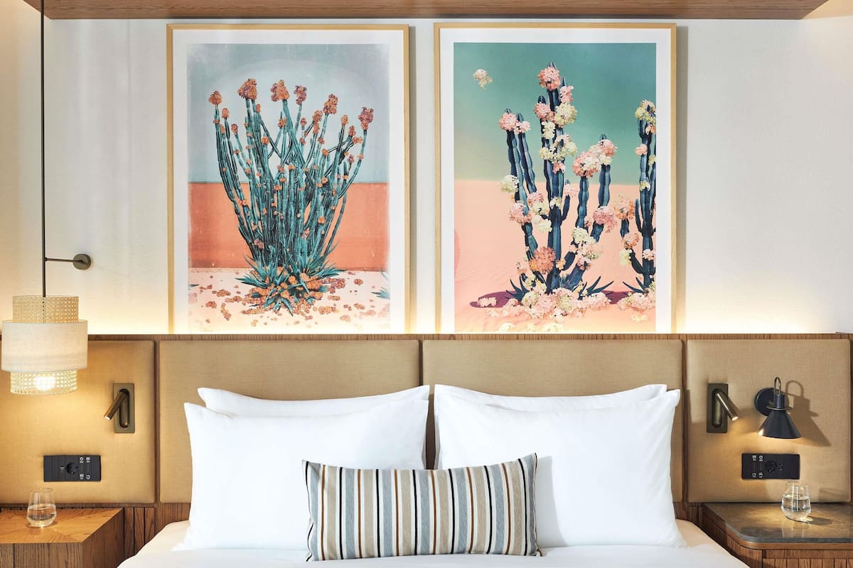 Quarto de hotel com uma cama, duas pinturas emolduradas, uma luminária de cabeceira e uma mesa de cabeceira com um copo de água.