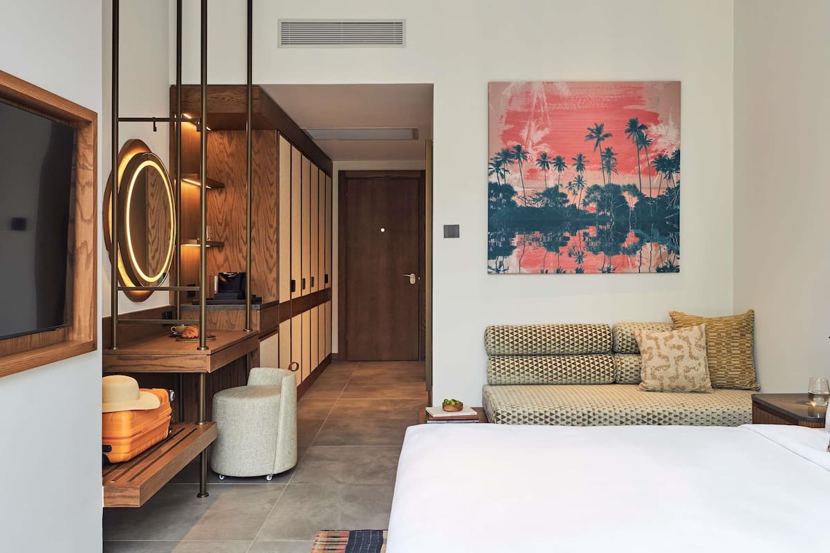 Quarto de hotel moderno com uma cama, um sofá, uma estante de madeira com malas, uma TV fixada na parede e uma pintura com tema tropical.