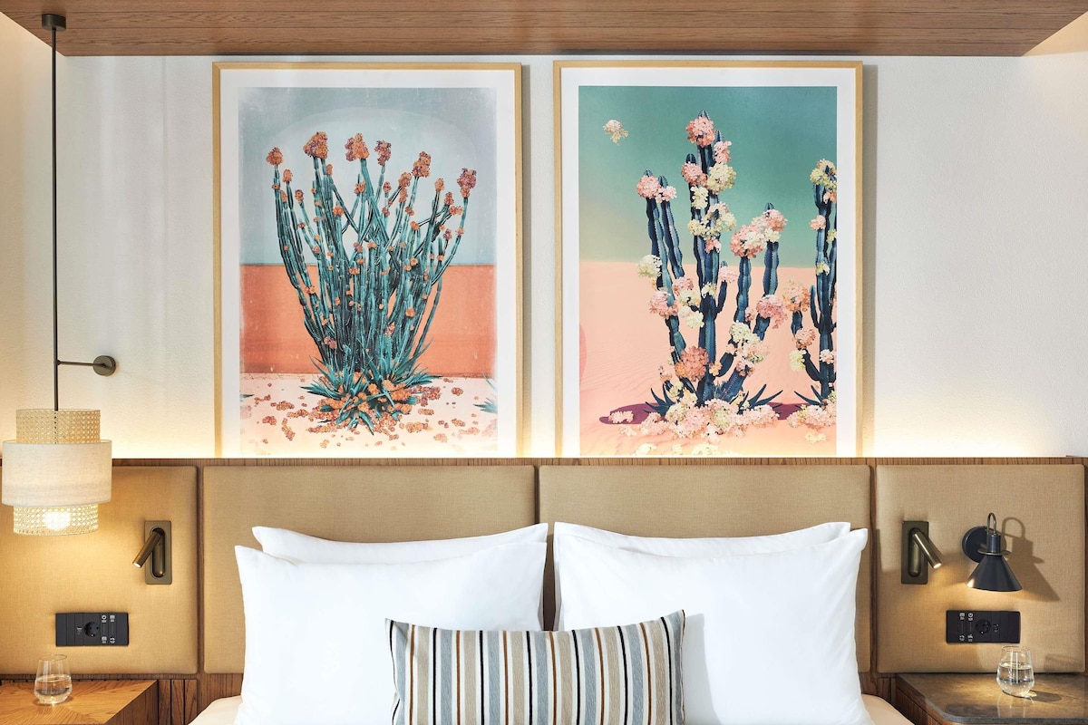Une chambre d’hôtel avec un lit, deux tableaux encadrés, une lampe de chevet et une table de chevet sur laquelle se trouve un verre d’eau.