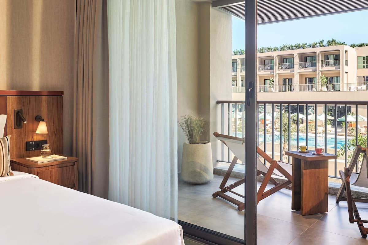 Quarto de hotel com cama, mesa de cabeceira com luminária, janela grande com cortinas leves e vista para uma piscina e edifícios.