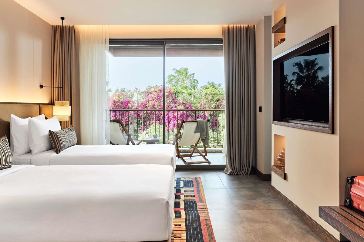 Une chambre d’hôtel avec un lit, une chaise, une télévision et un balcon donnant sur des fleurs roses.
