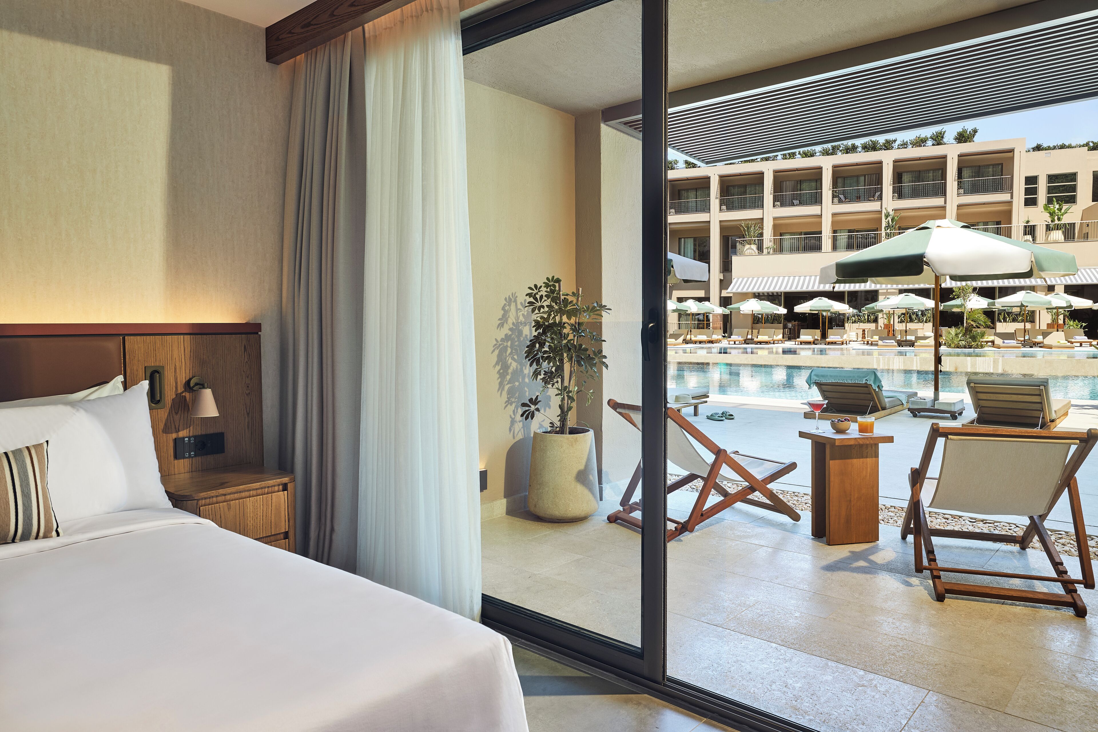 Une chambre d’hôtel avec un grand lit, une table de chevet, une lampe et une vue sur l’espace piscine, avec des chaises longues et des parasols.