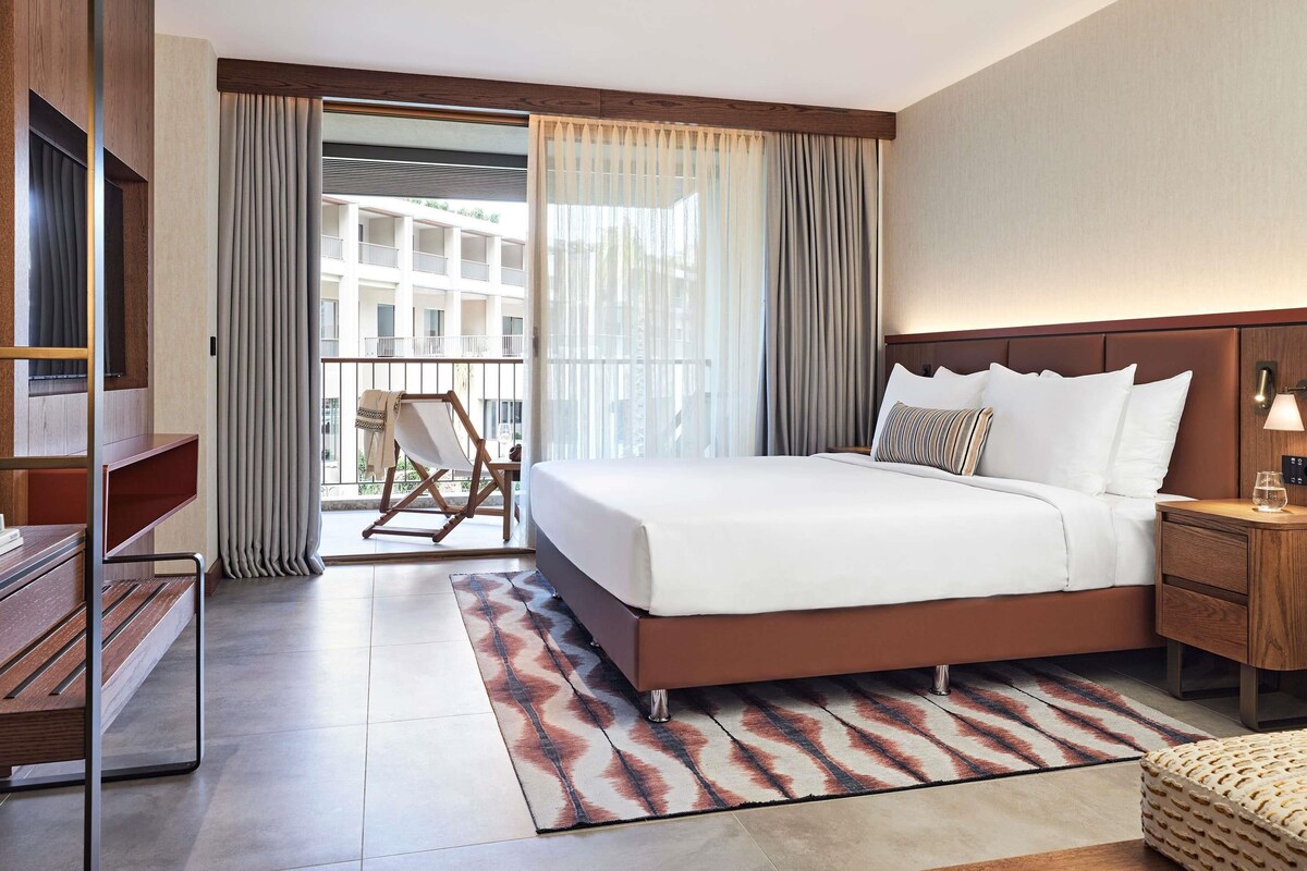 Une chambre d’hôtel avec un grand lit, une table de chevet en bois, un bureau, une chaise et une vue sur un immeuble par la fenêtre.