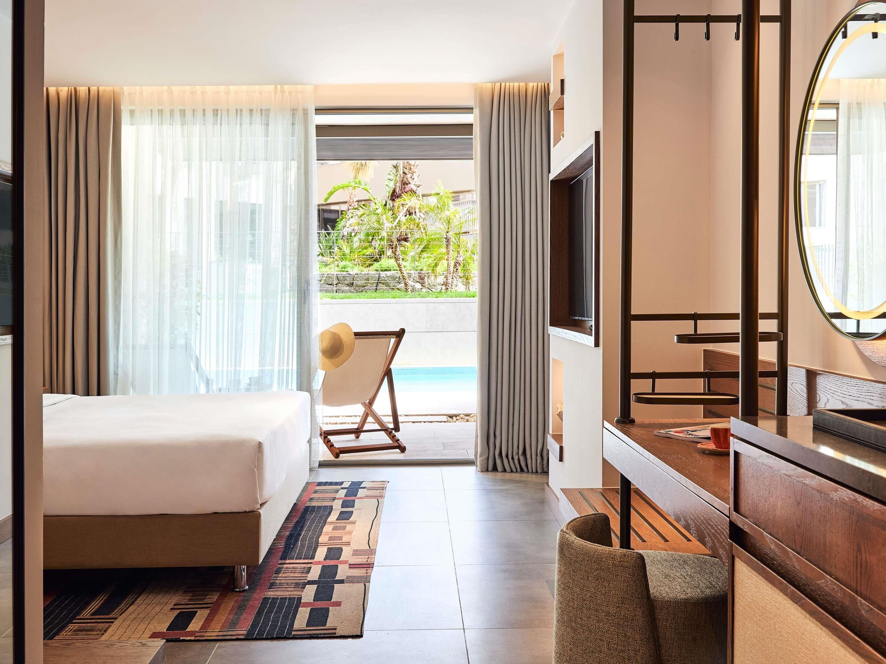 A hotel room with a bed, a TV, a desk, and a view of a pool and greenery.