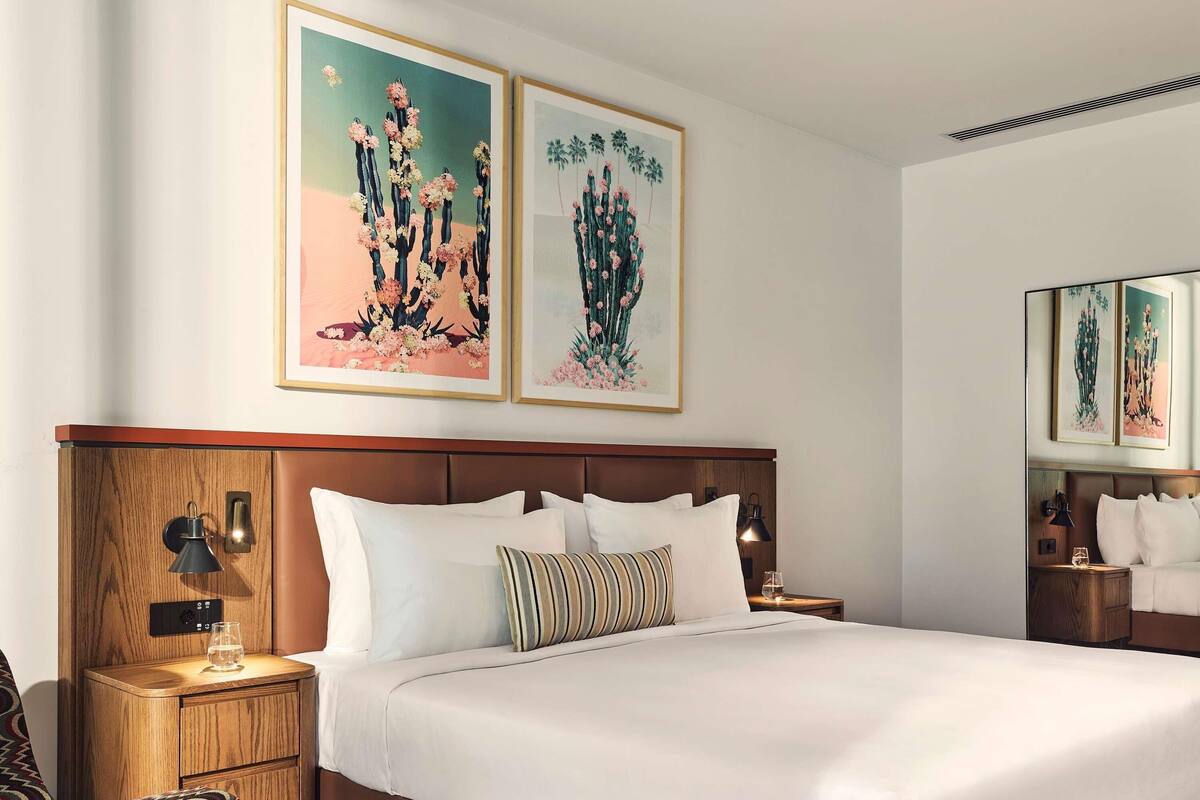 Quarto de hotel com uma cama grande, cabeceira de madeira, duas mesas de cabeceira com abajures e obras de arte nas paredes.