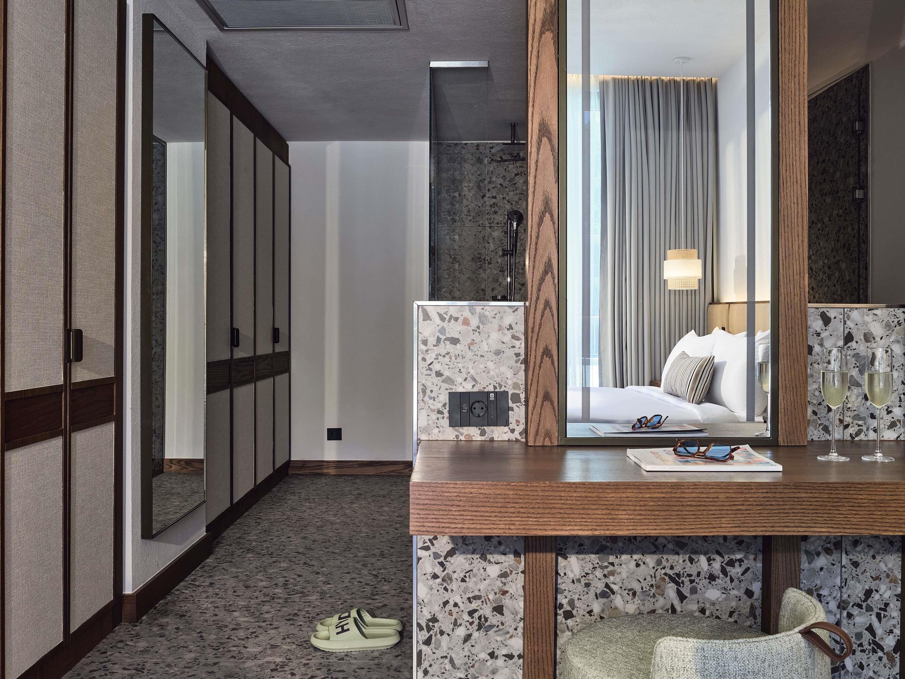 Ein Hotelzimmer mit einem großen Spiegel, einem Bett mit weißer Bettwäsche, einem Holztisch mit einem Laptop und einem Paar Hausschuhen auf dem Boden.