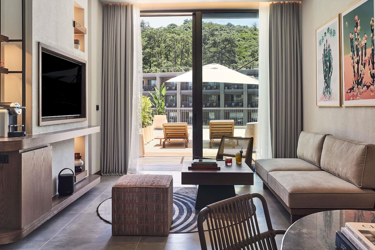 Un salon moderne doté d’un téléviseur à écran plat, d’un canapé, d’une table basse et offrant une vue sur un balcon aménagé avec du mobilier d’extérieur.