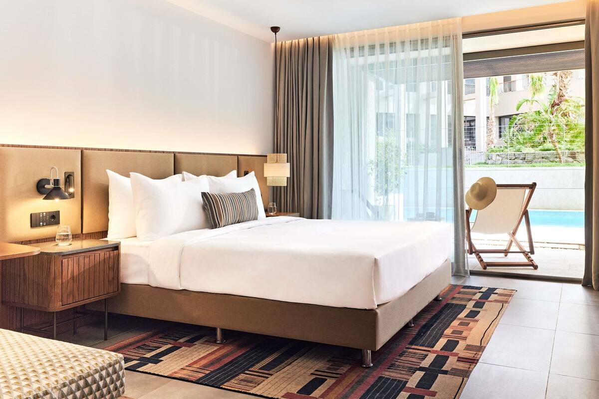 Quarto de hotel com uma cama grande, uma mesa de cabeceira com abajur, uma cadeira junto à janela e vista para uma piscina.