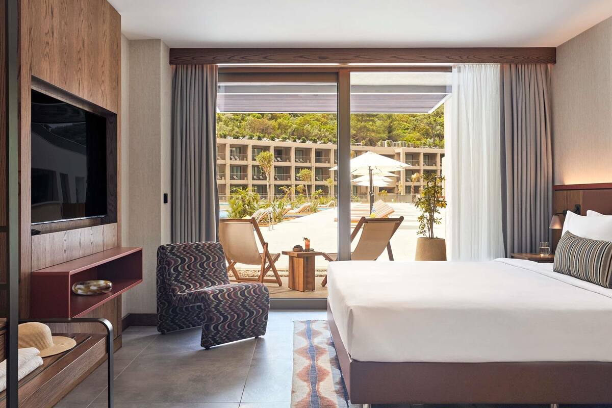 Quarto de hotel com uma cama grande, televisão presa a um painel de madeira, área de estar com uma cadeira e uma mesinha, e vista para uma piscina e um complexo de edifícios.