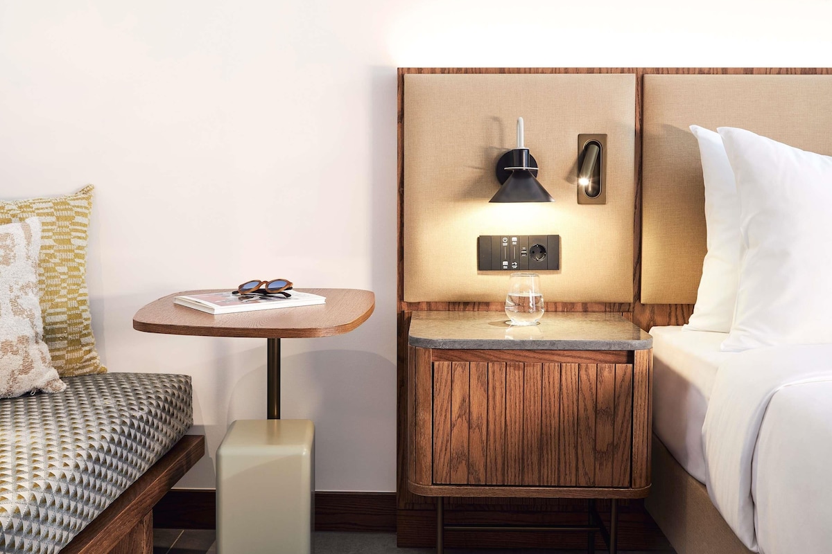 A hotel room with a bed, a bedside table with a lamp, a small table with a glass of water, and a pillow.