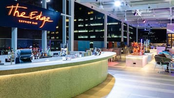 A bar area with a neon sign, bar counter, seating, and a view of the cityscape.