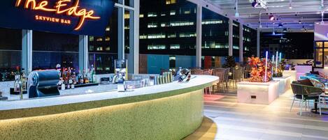 A bar area with a neon sign, bar counter, seating, and a view of the cityscape.