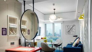 Une chambre d’hôtel moderne dotée d’un grand miroir, d’un bureau avec un plateau rouge, d’un canapé bleu, d’un coin repas offrant une vue sur la ville et d’une grande fenêtre.