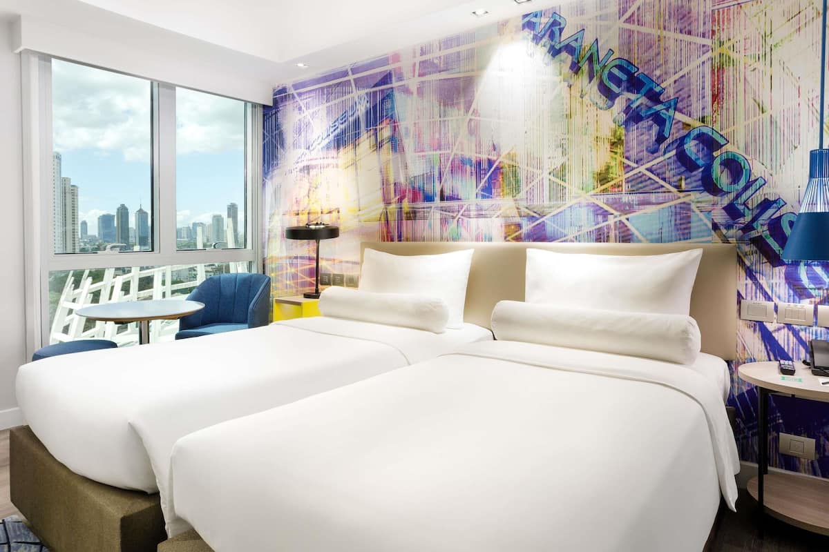 Ein Hotelzimmer mit zwei Betten, Blick auf die Stadt, einer farbenfrohen abstrakten Wand und einem kleinen Tisch mit einer Lampe.